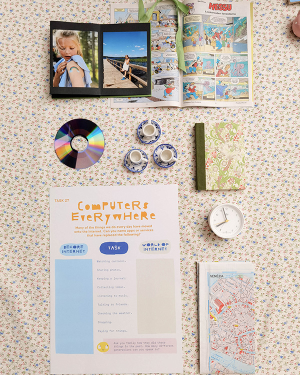 Flatlay showing Hello Ruby activity page about Computers Everywhere with photo album, comic book, notebooks, teacups, and clock on floral patterned surface